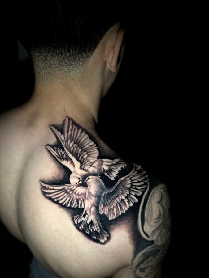 Two doves in flight tattooed in black and grey realism across a shoulder
