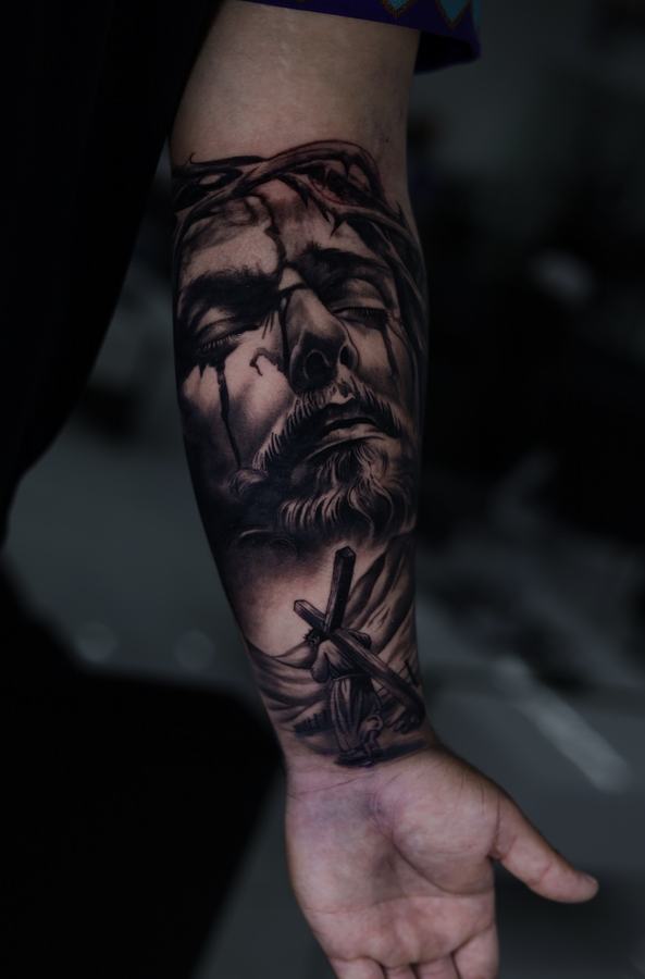Jesus with the crown of thorns above a scene of the cross, black and grey realism forearm tattoo