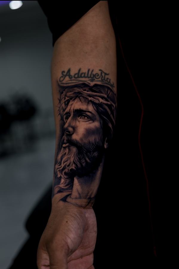 Jesus wearing the crown of thorns with script lettering above, black and grey realism forearm tattoo