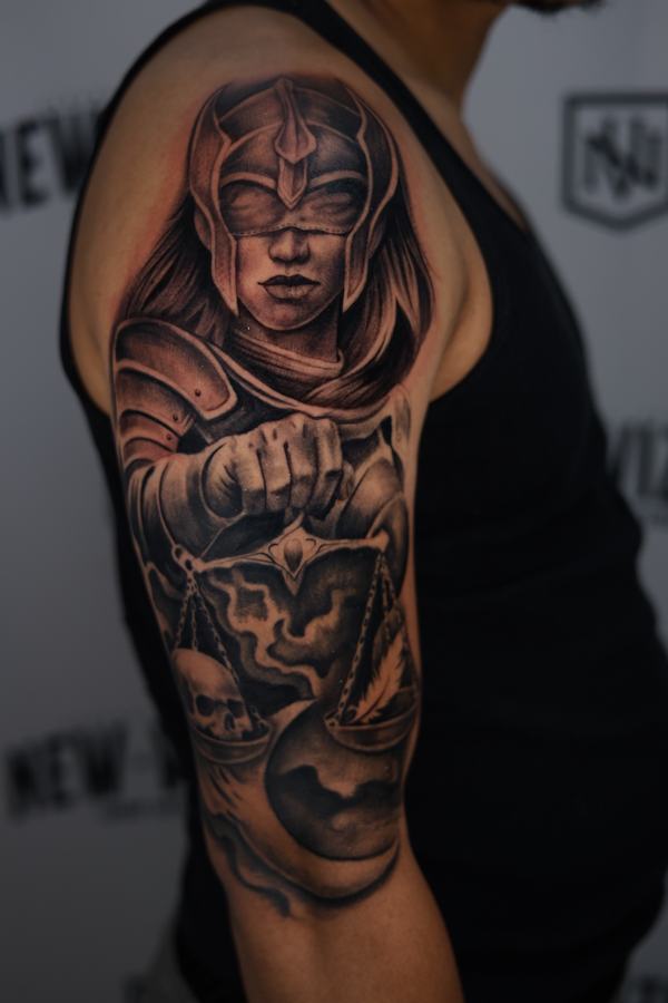 Blindfolded Lady Justice in armour holding scales, black and grey realism tattoo on an upper arm