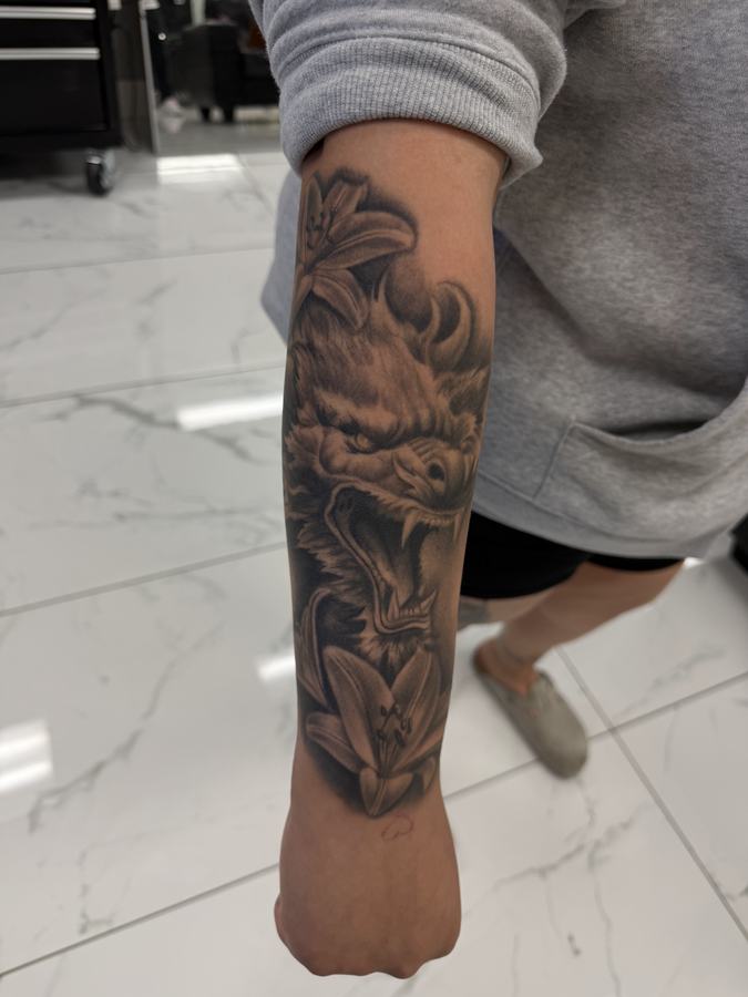 Lion tattoo surrounded by lilies in black and grey realism on a forearm sleeve