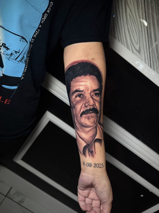 Memorial portrait tattoo of a man with a moustache and a date beneath, black and grey realism on a forearm