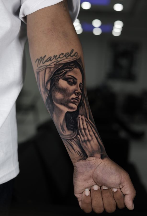Virgin Mary with praying hands and script name, black and grey realism tattoo on a forearm