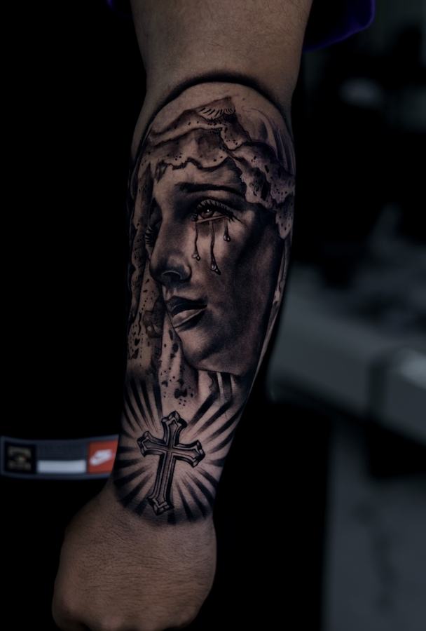 Weeping Virgin Mary above a cross, black and grey realism tattoo on a forearm